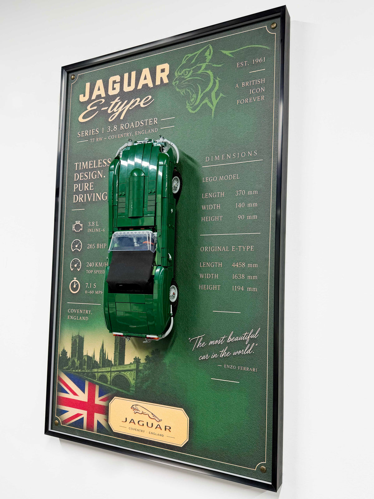 A framed vintage Jaguar E-type car display mounted on a white wall. The display features a green die-cast model car suspended over printed poster artwork with Jaguar branding, dimensions, and a small Union Jack flag.