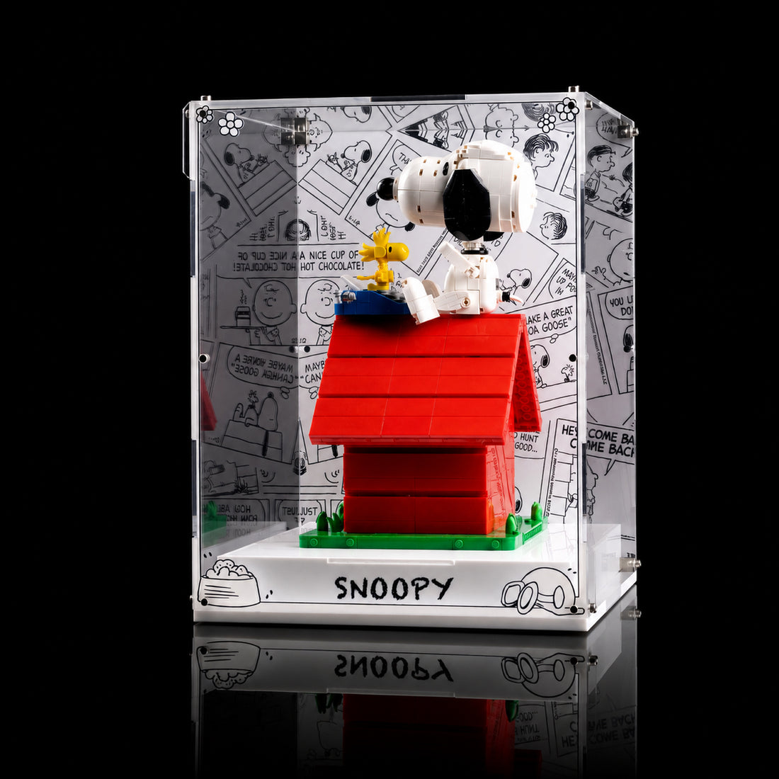 LEGO Ideas Peanuts: Snoopy's Doghouse acrylic display case (21368) shown with the assembled toy inside. The clear rectangular case features a white printed background with black comic-style drawings and the word 'SNOOPY' at the bottom.