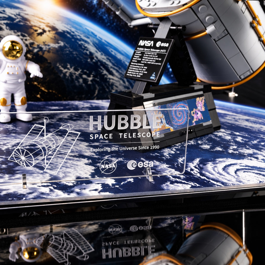 LEGO Hubble Space Telescope acrylic display case with astronaut figures, shown on a dark space-themed background. The case contains a detailed model of the Hubble telescope with Earth visible through the printed outer display panel.